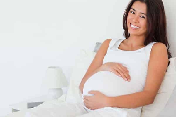 Women laying in bed holding her belly while pregnant with a smile.