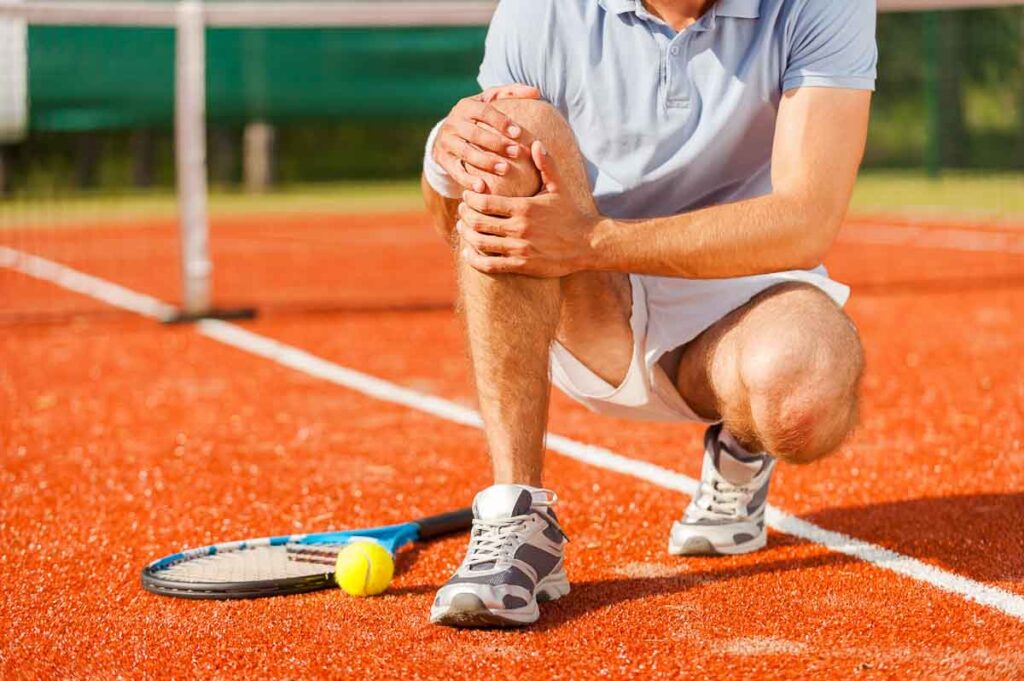 Man on tennis court, holding his knee in pain from a sports injury.